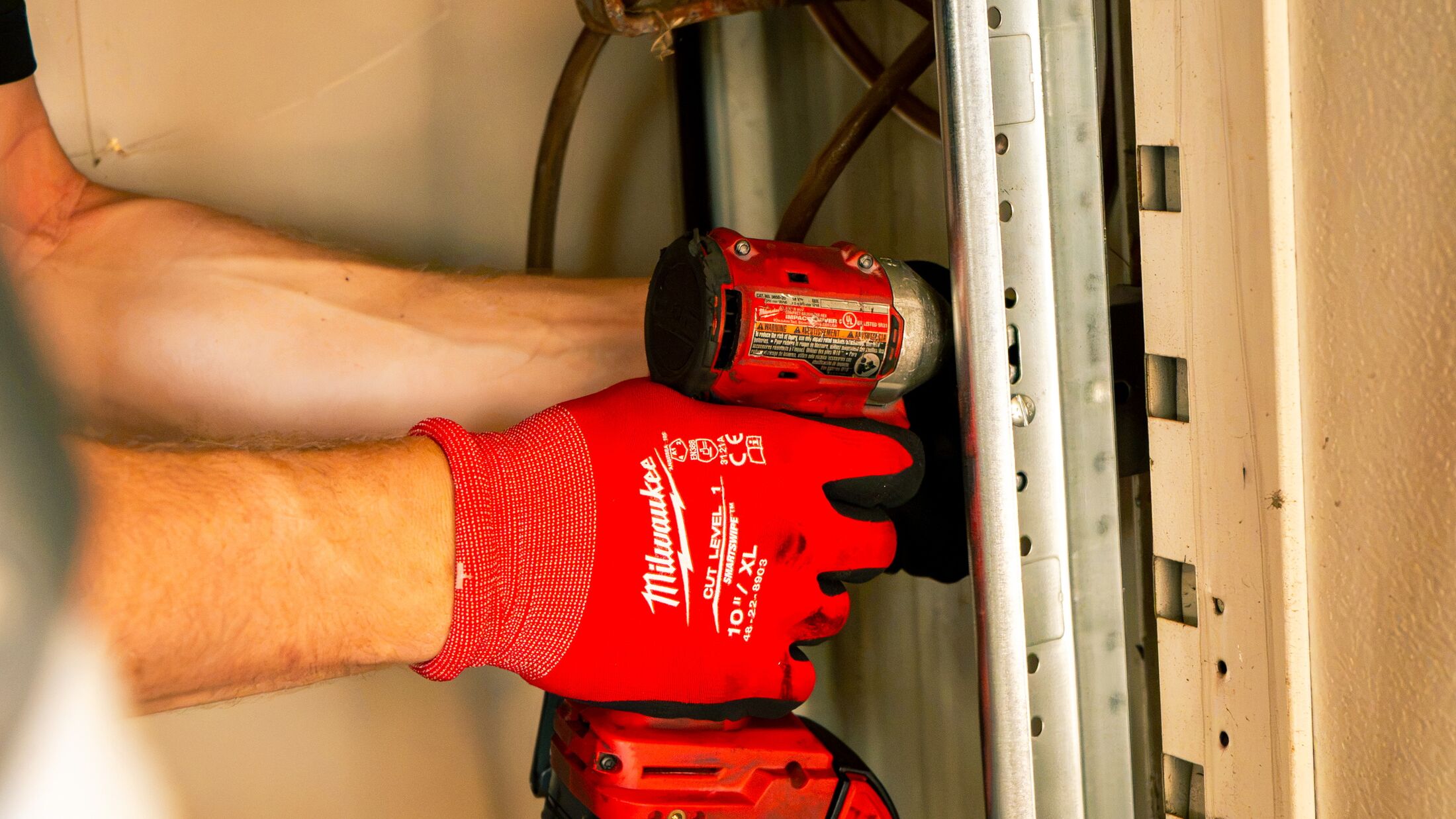 Close-up of a gloved hand and impact driver at a garage door's vertical track — worn track and rollers are what actually gives way in high wind